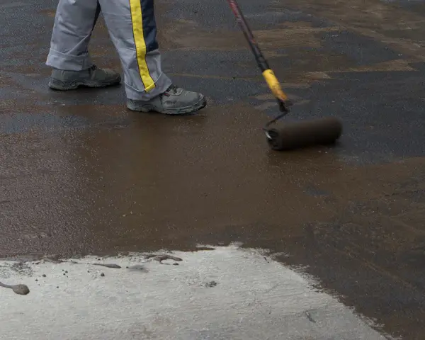 Pracownik wykonuje hydroizolację chudziaka, nakładając masę bitumiczną wałkiem.
