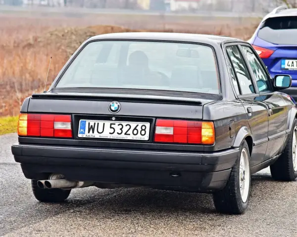 Tył czarnego BMW E30 z tablicą rejestracyjną WU 53268, holowanego przez niebieskiego SUV-a.