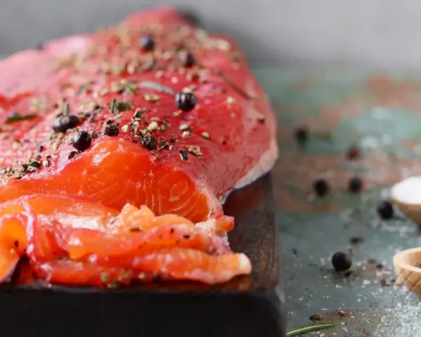 Un filet de gravlax de truite, assaisonné de poivre et d'herbes, prêt à être dégusté. Deux cuillères en bois contiennent sel et genièvre.