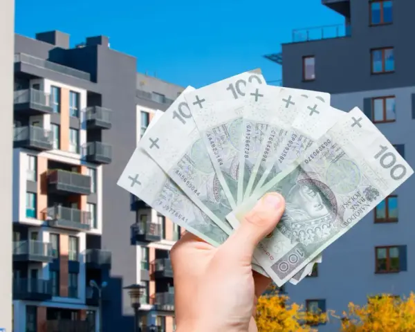 Dłoń trzyma banknoty 100 zł na tle nowoczesnych budynków mieszkalnych. Symbolizuje to podatek od sprzedaży nieruchomości.