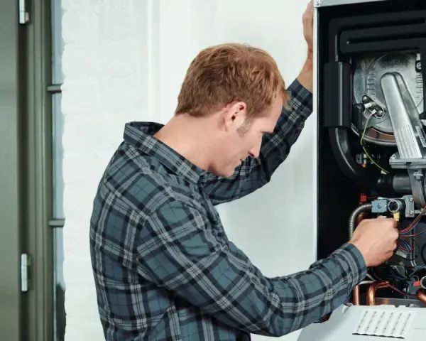 Tecnico che controlla lo scambiatore di calore della caldaia, con cavi e tubi visibili all'interno.