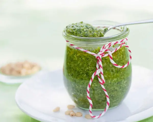 Un pot de pesto vert vif, décoré d'une ficelle rouge et blanche, prêt pour une recette pesto traditionnel. Quelques pignons de pin à côté.