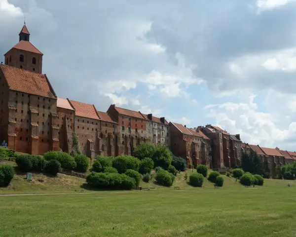 Długi, ceglany zamek w Grudziądzu w jeden dzień. Zielona trawa i krzewy przed zabytkową budowlą.