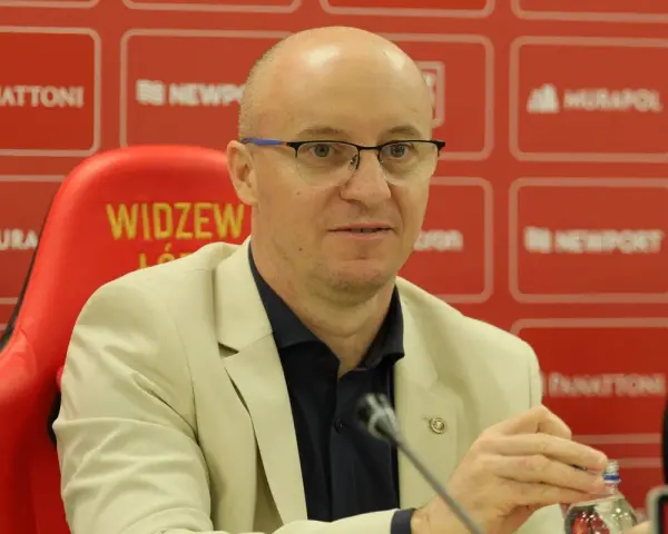Nowy trener Widzewa w jasnej marynarce, siedzi na czerwonym fotelu z logo klubu.