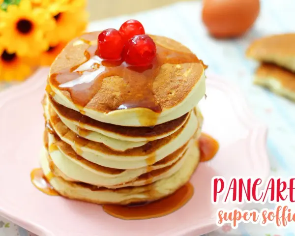 Pancakes con albumi, soffici e dorati, guarniti con sciroppo e ciliegie rosse, su un piatto rosa.