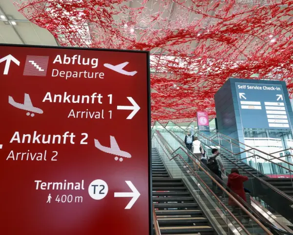 Czerwony znak z kierunkami na lotnisku w Niemczech: odloty, przyloty, terminal T2. W tle schody ruchome i instalacja artystyczna.