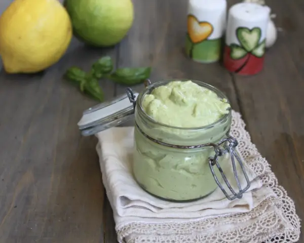 Un vasetto di cremoso pesto al limone, con limoni e basilico sullo sfondo.