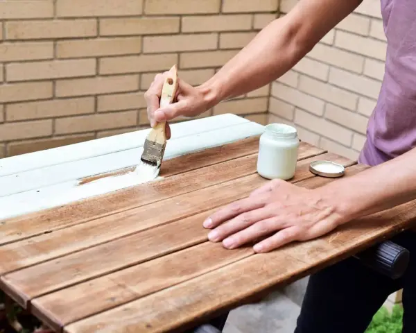 Persona pinta madera de blanco con brocha. Un frasco de pintura blanca y su tapa están cerca.