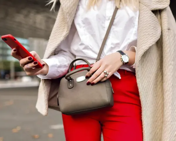 Une femme élégante, avec un manteau beige, un chemisier blanc et un pantalon rouge vif, tient un téléphone rouge et un petit sac gris. Quel entoilage pour sac choisir pour un tel look ?