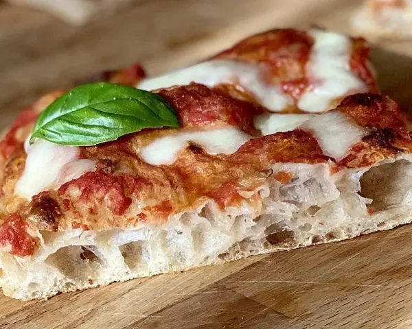 Fetta di pizza fatta in casa teglia, con crosta alveolata, pomodoro, mozzarella filante e una foglia di basilico fresco.
