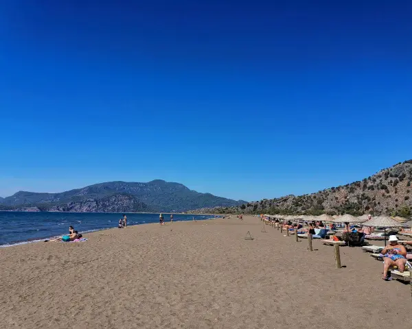 Słoneczna plaża w Sarigerme, z turkusowym morzem, górami w tle i rzędami leżaków pod słomianymi parasolami.