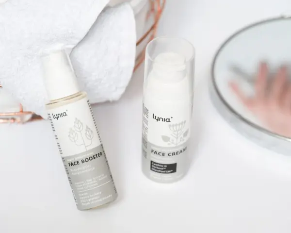 Białe opakowania kosmetyków Lynia: Face Booster i Face Cream, z widocznym składnikiem papaina. Obok ręczniki i lustro.