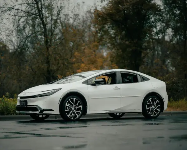 Biała Toyota Prius zaparkowana na mokrej drodze, z drzewami w tle.