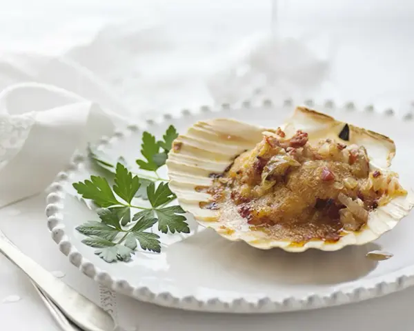 Vieiras gratinadas con tocino y perejil, servidas en su concha sobre un plato blanco.
