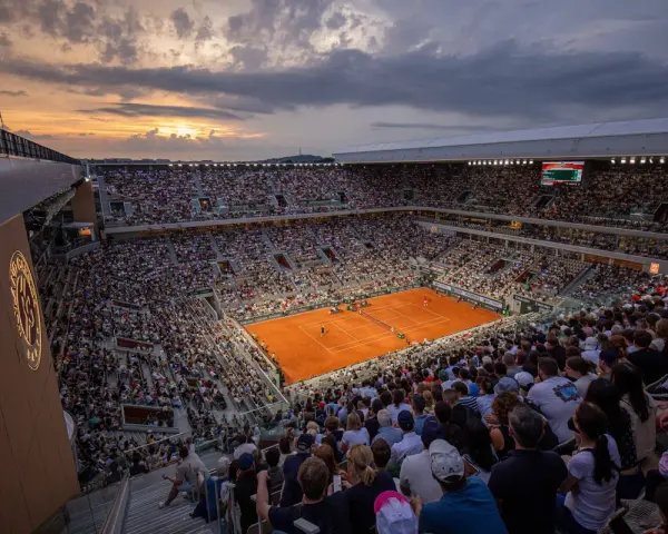 Widok na pełny stadion podczas meczu na kortach ziemnych. Zachodzące słońce oświetla trybuny pełne widzów, tworząc atmosferę emocjonującego **Roland Garros live**.