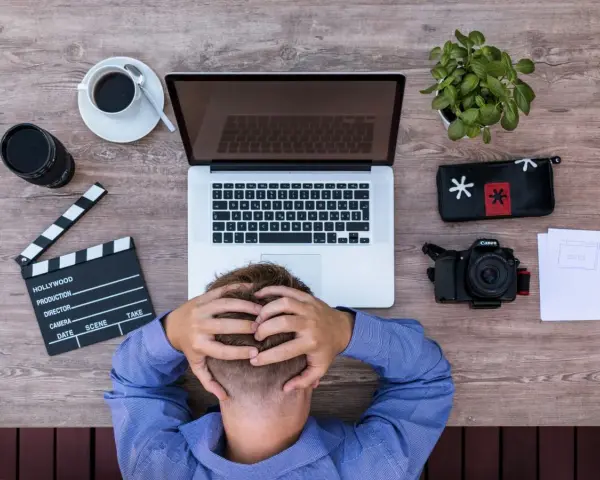 Frustracja twórcy filmowego. Na drewnianym biurku leży laptop, aparat, klaps, kawa i roślina.