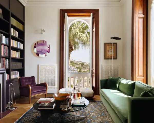 Soggiorno elegante in una casa stile Liberty, con divano verde, poltrona viola, libreria e vista panoramica.