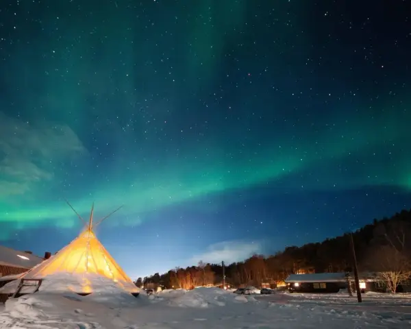 Nocne niebo nad ośnieżoną wioską, gdzie nie zachodzi słońce. Tańcząca zorza polarna nad oświetlonym tipi i domkami.