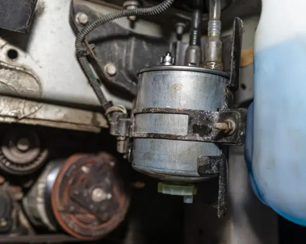 Kraftstoffpumpe defekt weiterfahren? Nicht ratsam. Nahaufnahme eines Kraftstofffilters im Motorraum, neben einem blauen Behälter.