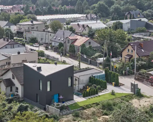Nowoczesny, czarny dom o trójkątnym kształcie wyróżnia się na tle tradycyjnych terenów mieszkaniowych b. Otoczony zielenią, z niebieskimi drzwiami.