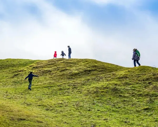 Famiglia esplora sentieri per bambini su una collina verdeggiante sotto un cielo azzurro e nuvoloso.