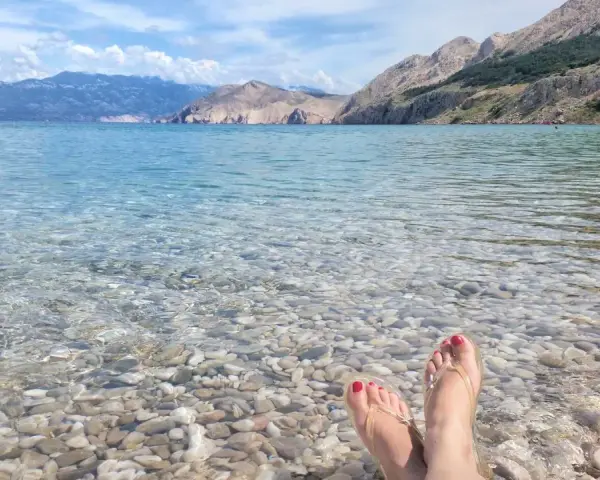 Stopy w klapkach na kamienistej plaży w **Krk National Park**. Woda krystalicznie czysta, w tle góry.