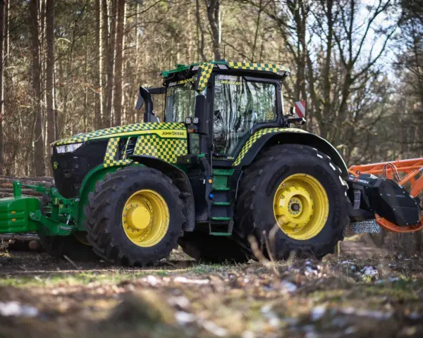 Zielony traktor John Deere z pomarańczowym mulczerem, gotowy do pracy w lesie.