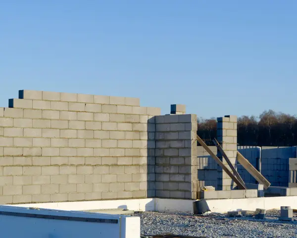 Budowa domu w toku. Wzniesiona ściana nośna z bloczków betonowych, w tle kolejne elementy konstrukcji i las.