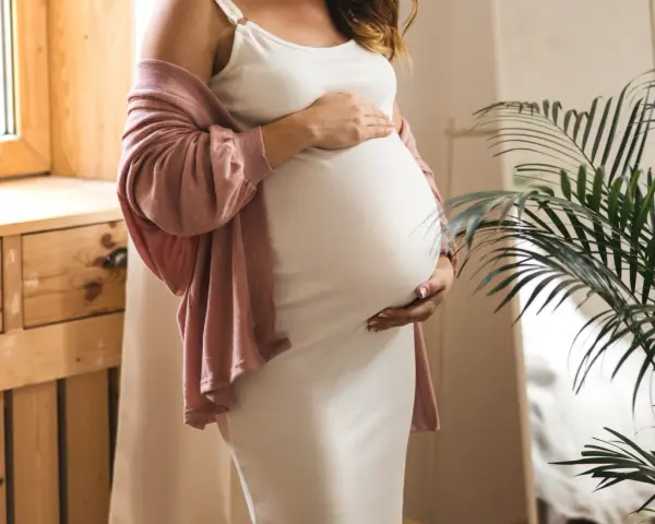 Frau in weißem Kleid und rosafarbenem Cardigan hält ihren Bauch, 34 ssw. Ein Fenster und eine Pflanze sind im Hintergrund.