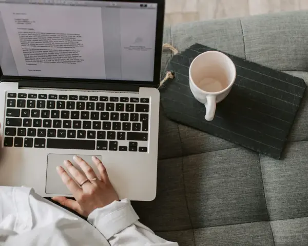 Person arbetar på laptop i soffan, en kopp står bredvid. Perfekt för dig som söker bra utbildningar på distans.