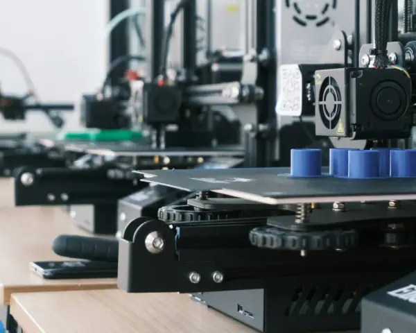 Drukarki 3D pracują, tworząc niebieskie cylindry. W tle widać kolejne maszyny, a na biurku leży smartfon, gotowy do zarządzania drukiem przez Raspberry Pi z OctoPrint.