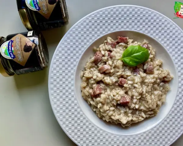 Aksamitne risotto z kawałkami kiełbasy, udekorowane listkiem bazylii. Obok stoją słoiczki z sosem truflowym, sugerujące jego obecność w daniu.
