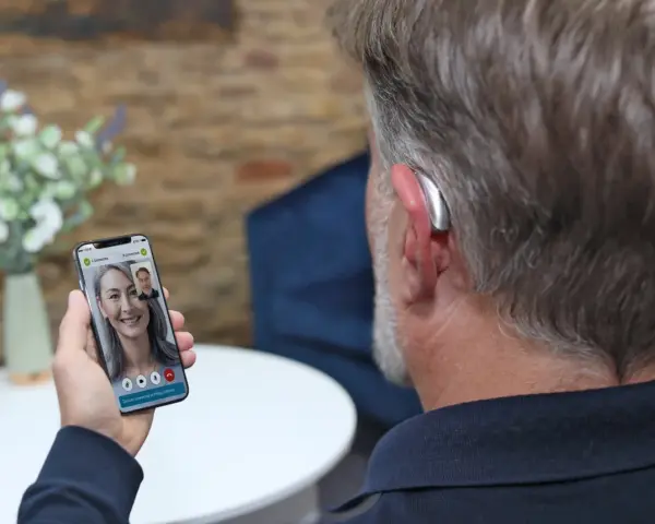 Mężczyzna z aparatem słuchowym rozmawia przez wideorozmowę na telefonie. Aplikacja na telefon pozwala na zdalne dopasowanie aparatów.