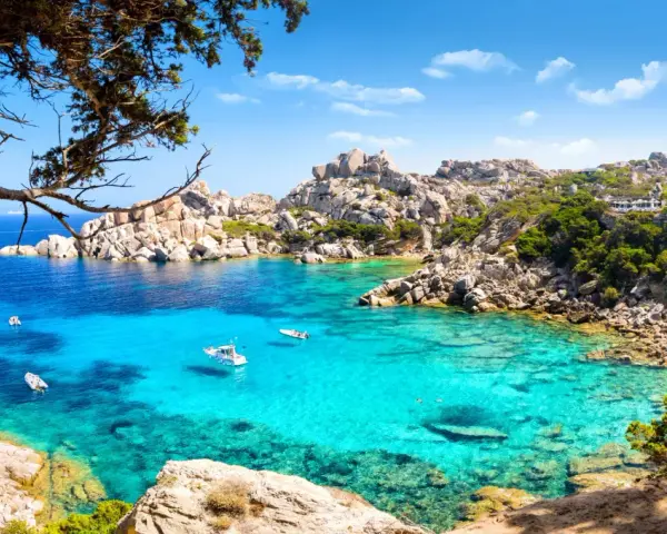 Turkusowa woda i skaliste wybrzeże Costa Smeralda na Sardynii. Kilka łodzi kołysze się na falach.