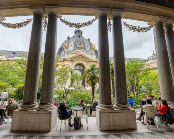 Vue sur le Grand Palais depuis un portique, avec des visiteurs profitant d'un café dans le jardin. Un petit palais, un grand palais.