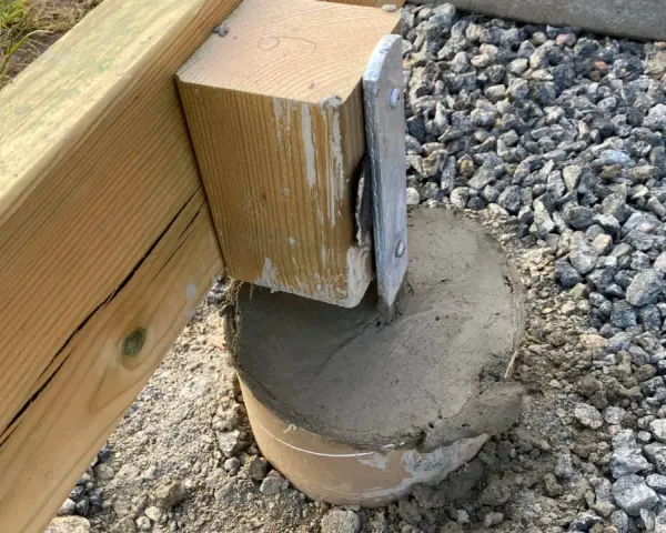 En trästolpe fästs i en betongplint, en del av arbetet med att bygga altan på plintar. Grus och jord syns runt plinten.