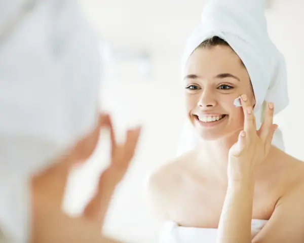 Una mujer sonriente con toalla en la cabeza se aplica crema en la cara, un paso esencial para como cuidar la piel de la cara.