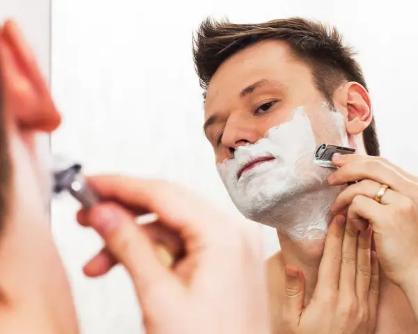 Hombre con espuma de afeitar en la cara, afeitándose con una maquinilla. ¿Es mejor afeitarse antes o después de la ducha?