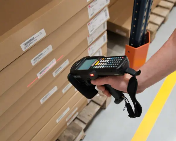 Mano escaneando cajas apiladas con un dispositivo, usando la **fórmula de Wilson** para optimizar el inventario.