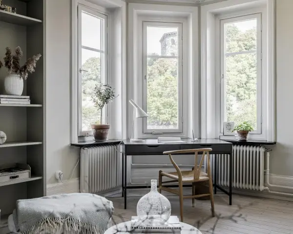 Una **scrivania sotto finestra** con vista sul verde. Un angolo studio accogliente con piante, lampada e una sedia di design.