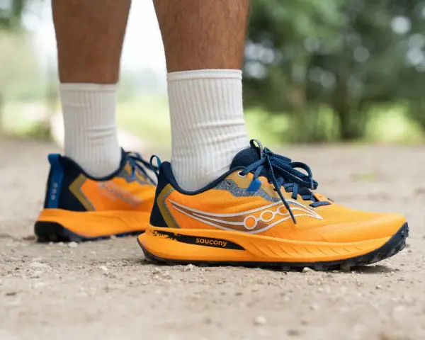 Gambe di persona che indossa scarpe da trail running Saucony arancioni e blu su un sentiero sterrato.