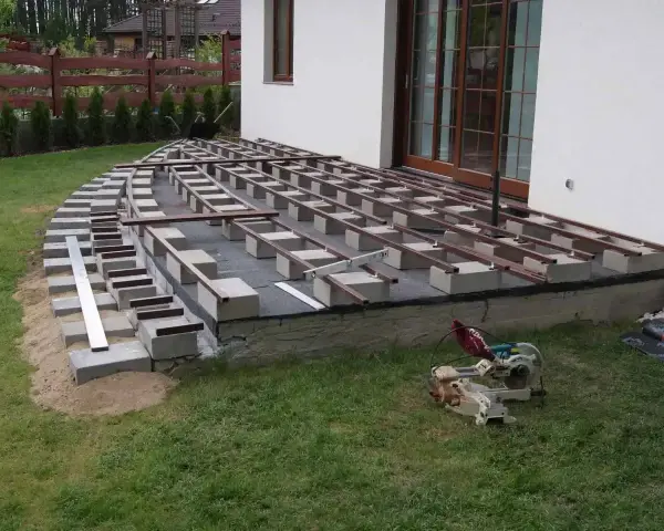 Budowa tarasu z deski kompozytowej w toku. Widać konstrukcję nośną z bloczków betonowych i legarów, gotową do ułożenia desek.