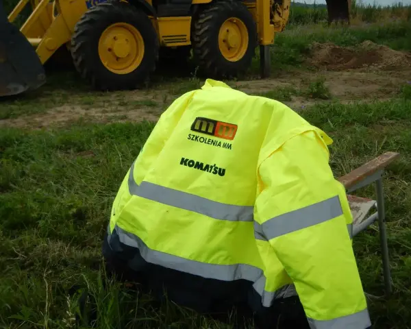 Żółta kamizelka z logo Komatsu i napisem "Szkolenia MM" leży na krześle. To przygotowanie do egzaminu na ładowarkę, gdzie pytania i odpowiedzi są kluczowe.