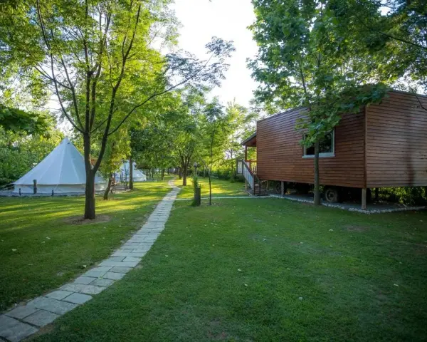 Malowniczy camping w Albanii z białymi namiotami typu bell i drewnianym domkiem na palach, otoczony zielenią.