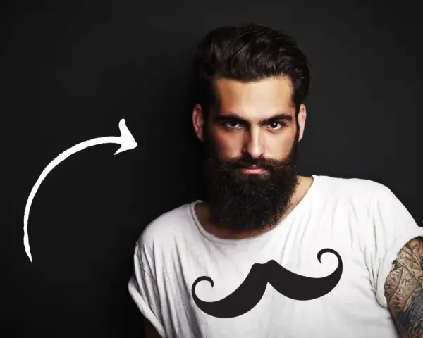 Hombre con barba tupida, quizás decidiendo dejarse barba por primera vez. Camiseta blanca con estampado de bigote.