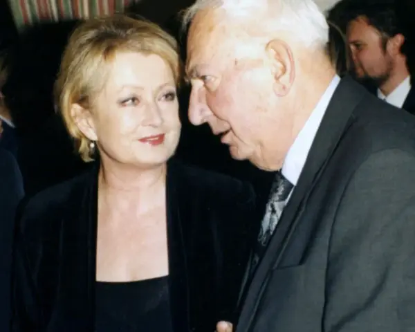 Ewa Holoubek w czarnej sukni i marynarce rozmawia z mężczyzną w garniturze.