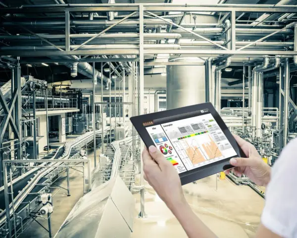 Operator kontroluje linię produkcyjną za pomocą tabletu, na którym widnieje wykres zużycia energii. W tle widać skomplikowany system rur i taśmociągów. Czujnik podczerwieni może być kluczowy dla monitorowania procesów.