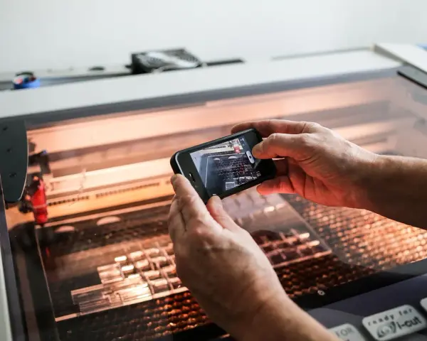 Hands hold a phone, capturing a laser cutter working on intricate acrylic projects.