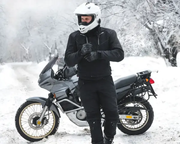 Motociclista con casco bianco e abbigliamento invernale nero, pronto per un'avventura in moto inverno su una strada innevata.
