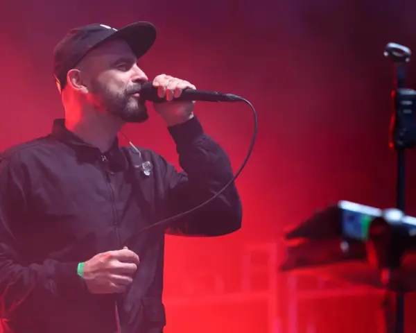 Wokalista z brodą i czapką na głowie śpiewa do mikrofonu na scenie. Czerwone światła tworzą atmosferę koncertu. Tauron Nowa Muzyka line-up.